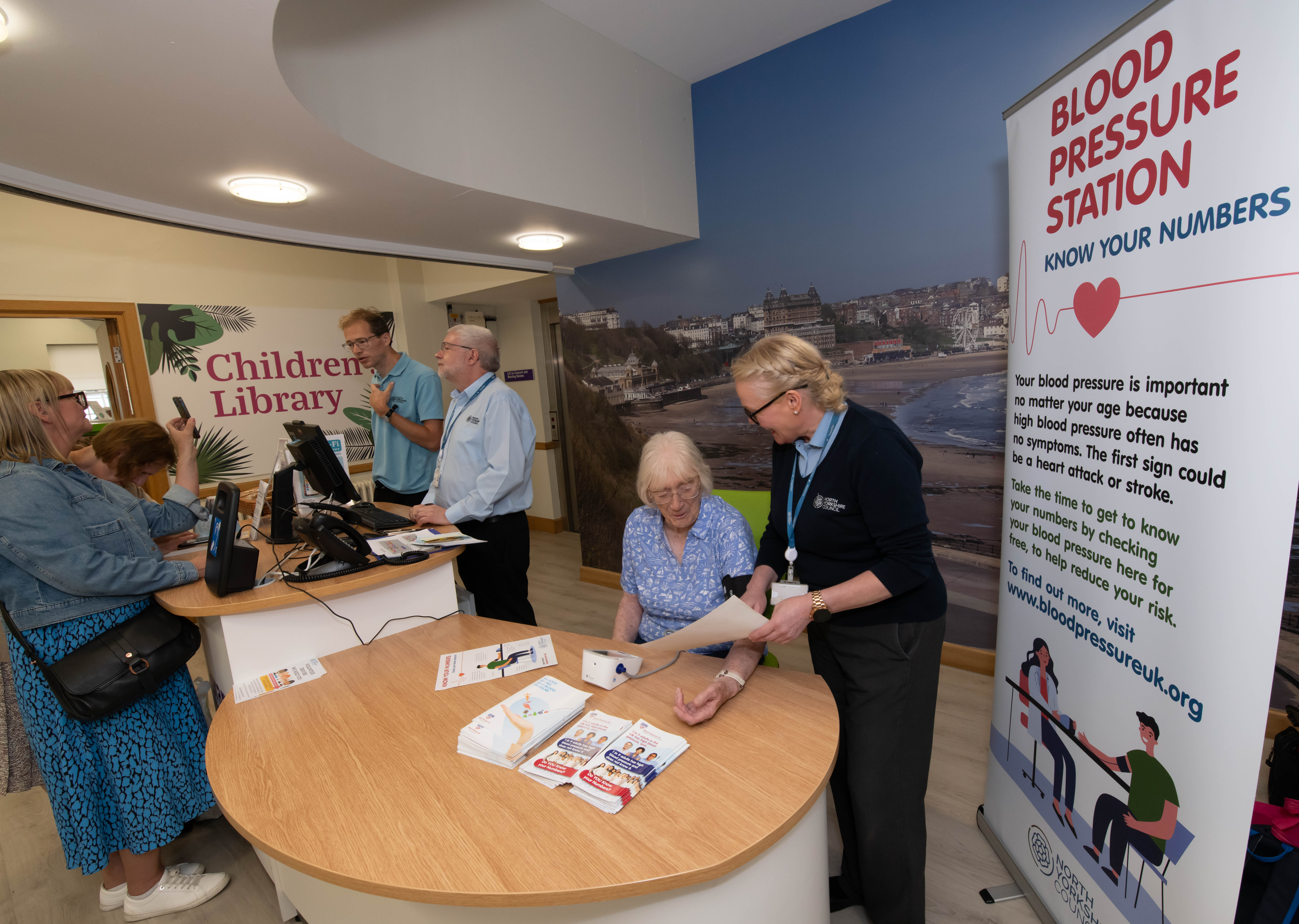 Image showing a person having their blood pressure taken happily chatting with a staff member whilst a group of people chat in the background.